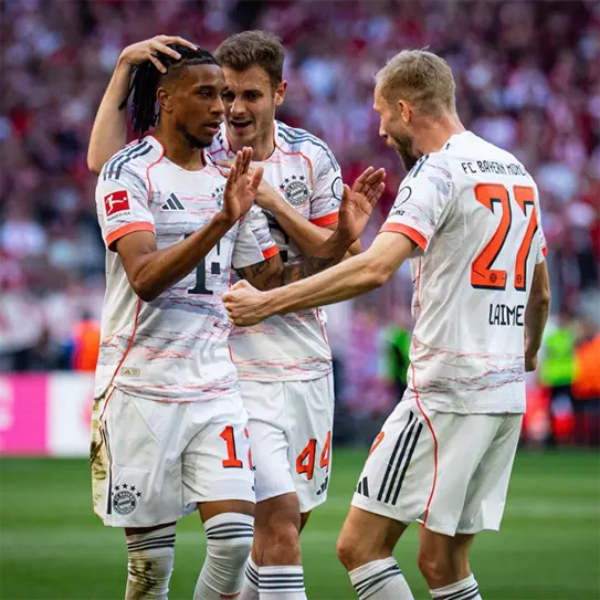 Three soccer players from Bayern Munich celebrate a goal on the field. One player, in a white jersey with red accents, is being congratulated by two teammates. The atmosphere is lively, with fans visible in the background, showcasing excitement and team spirit.