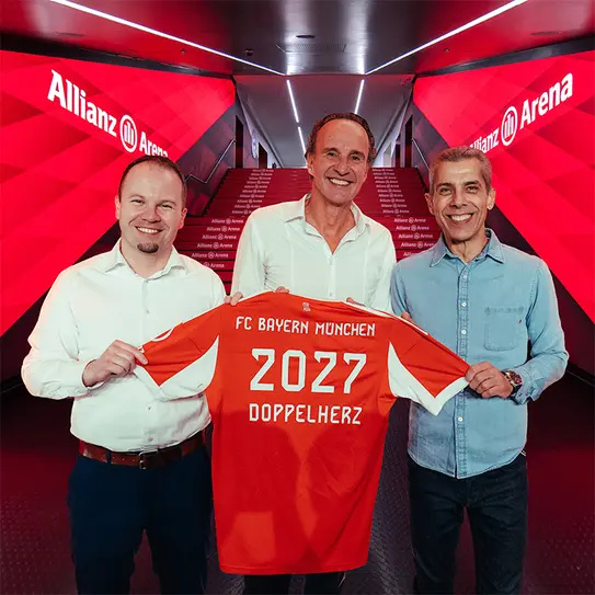 Three men stand in a brightly lit hallway at Allianz Arena, holding an orange FC Bayern München jersey. The jersey features the numbers "2027" and the word "Doppelherz." The background has vibrant red graphics, highlighting the team's branding.