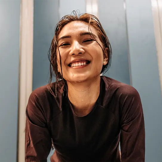 A smiling young woman with wet hair is leaning forward, expressing joy and enthusiasm. She is wearing a dark athletic top, and the background features light-colored, textured panels, enhancing her positive energy.