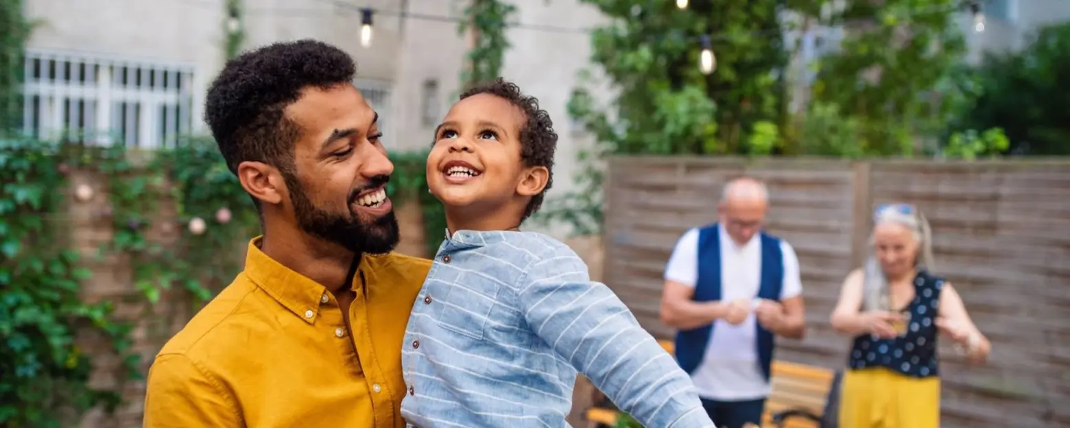 A joyful scene of a father holding his smiling son outdoors, surrounded by greenery. The father, wearing a yellow shirt, looks down at his son, dressed in a blue striped shirt, who beams with happiness. In the background, other people engage casually in a social setting.