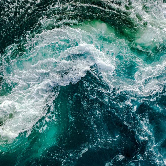 Luftaufnahme eines aufgewühlten Meeres mit weißen Schaumkronen und kreisenden Strömungen.